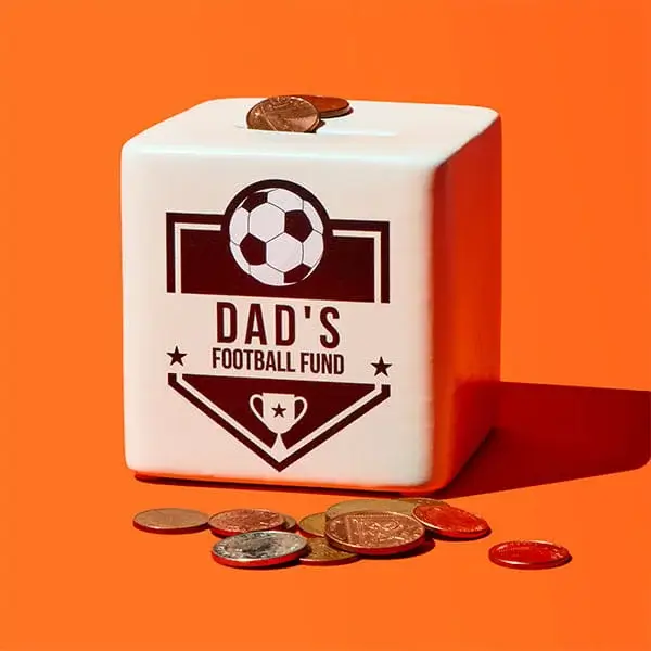 A white ceramic money box labelled Dad’s Football Fund with a football graphic, shown with coins on top and scattered at the base against a bright orange background.
